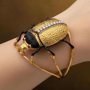 May include: A gold-toned bracelet featuring a detailed beetle design. The beetle has a textured gold body, black head and legs, and is adorned with small pearls. Red accents are visible on the beetle's head. The bracelet is a unique piece of jewelry.