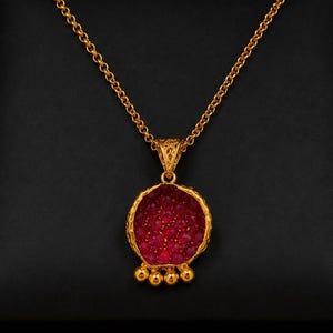 May include: A gold chain necklace featuring a circular pendant with a textured, deep red gemstone center. The pendant has a decorative filigree bail and four small gold spheres hanging from the bottom edge, creating a pomegranate-like shape.