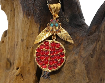 Peut inclure: Un collier en or avec un pendentif en forme de grenade. Le pendentif est en métal rouge et or et présente un design détaillé. Le collier est suspendu à un support en bois.