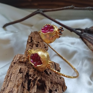May include: A gold bracelet featuring two pomegranate-shaped charms. The charms are filled with red gemstones and have black accents.