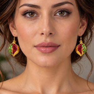 May include: A pair of gold-toned drop earrings shaped like pomegranates. Each earring features a textured gold base, a cluster of deep red crystals representing seeds, and small green enameled leaves attached to the top of the fruit design.