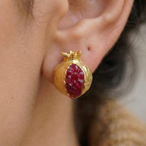 May include: Gold-colored stud earrings in the shape of pomegranates. Each earring features a textured gold exterior and a vibrant red interior, resembling the fruit's seeds. The earrings are worn on an ear.