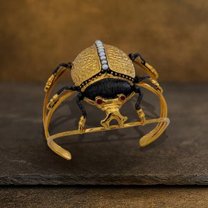 May include: Gold-toned cuff bracelet featuring a detailed scarab beetle design. The beetle has a textured gold body, black accents, and red gemstone eyes. A row of small white pearls adorns the top. The bracelet is set against a dark background.