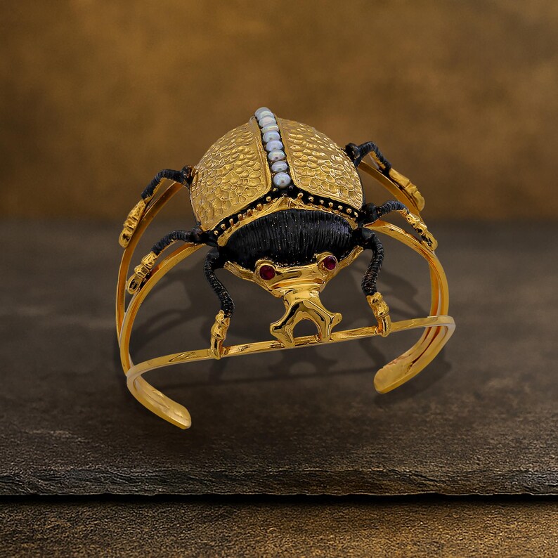 May include: Gold-toned cuff bracelet featuring a detailed scarab beetle design. The beetle has a textured gold body, black accents, and red gemstone eyes. A row of small white pearls adorns the top. The bracelet is set against a dark background.