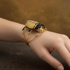 May include: Gold-toned cuff bracelet featuring a detailed beetle design. The beetle has a textured gold body, black head, and legs, with small pearl accents. The bracelet is worn on a wrist.