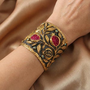 Peut inclure: Un large bracelet manchette orné d'un motif de grenade. Le bracelet est de couleur dorée avec des accents noirs et est orné de pierres précieuses rouges et turquoise. Le design comprend des feuilles et des grenades.