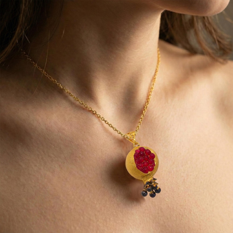May include: A gold-colored necklace featuring a pendant shaped like a pomegranate. The pendant has a textured gold exterior with a cluster of red stones representing the seeds. Small black beads hang from the bottom.