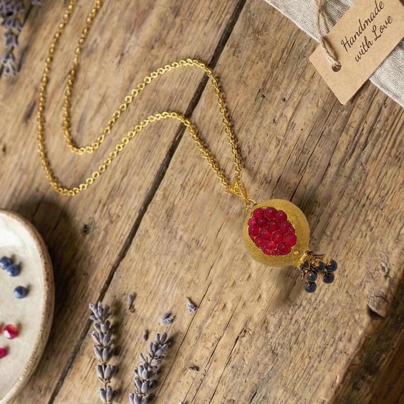 May include: A gold-colored necklace with a pendant shaped like a pomegranate. The pendant features a textured gold exterior and a cluster of red gems representing the seeds. Small blue gems are at the base.