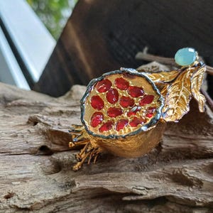 May include: A gold-toned pomegranate brooch with red glass stones and a green stone accent. The brooch is detailed with a gold leaf design.