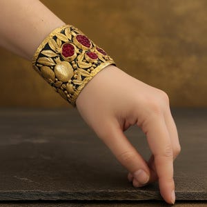 May include: A wide gold cuff bracelet with a black background, featuring a detailed design of pomegranates and leaves. The bracelet has red gemstone accents and is worn on a wrist.