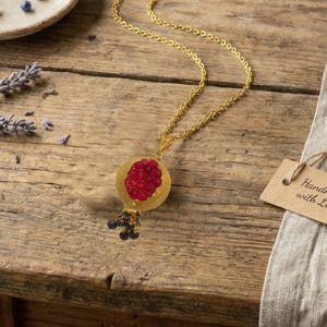 May include: A gold-colored necklace with a pendant shaped like a pomegranate. The pendant features a red, jewel-like interior and a cluster of dark blue beads at the bottom. The necklace is displayed on a wooden surface.