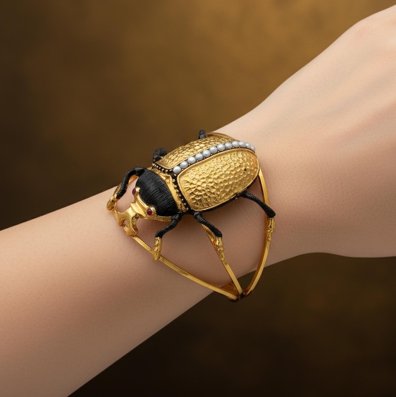 May include: A gold-toned bracelet featuring a detailed beetle design. The beetle has a textured gold body, black head and legs, and is adorned with small pearls. Red accents are visible on the beetle's head. The bracelet is a unique piece of jewelry.