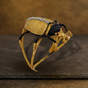May include: A gold-toned cuff bracelet featuring a detailed scarab beetle design. The beetle has a textured gold body, black accents, and a row of small white pearls. The bracelet includes red gemstone accents and is set against a dark background.