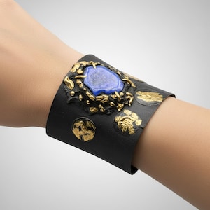 May include: A black cuff bracelet with gold accents and a large, oval, blue gemstone at the center. The bracelet features intricate gold detailing around the gemstone and on the surface.