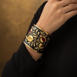 May include: Wide cuff bracelet with a black background and gold pomegranate design. The bracelet features gold leaves, pomegranates with red gems, and small turquoise accents. The bracelet is worn on a wrist, showcasing the intricate details and craftsmanship.