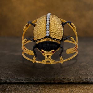 May include: A gold-toned cuff bracelet featuring a detailed scarab beetle design. The beetle has a textured gold shell, black body, and red gemstone eyes. A row of small pearls accents the shell. The bracelet has a unique, open design.