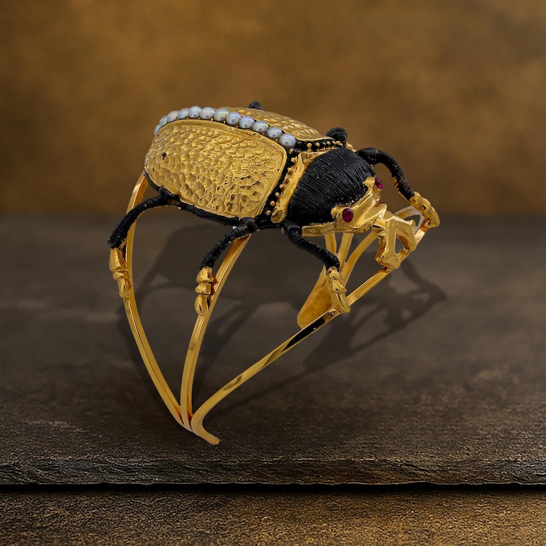 May include: A gold-toned bracelet featuring a detailed scarab beetle design. The beetle has a textured gold body, black head and legs, and is adorned with small pearls and red accents. The bracelet's frame is also gold, with an open, curved design.