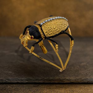 May include: A gold-toned beetle-shaped bracelet with intricate detailing. The beetle features a textured gold body, black head and legs, and small red accents. A row of small white pearls lines the top of the beetle's body. The bracelet has a hinged design.