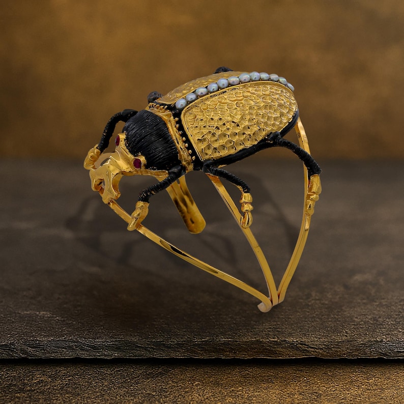 May include: A gold-toned beetle-shaped bracelet with intricate detailing. The beetle features a textured gold body, black head and legs, and small red accents. A row of small white pearls lines the top of the beetle's body. The bracelet has a hinged design.