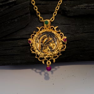 May include: Gold-toned pendant necklace featuring a detailed coin design with a profile of a person. The pendant is adorned with red and green gemstones and a dangling pink gemstone. The necklace has a delicate chain.