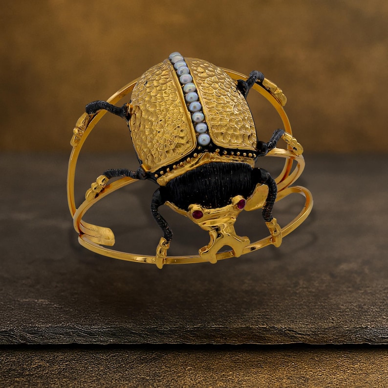 May include: A gold-toned bracelet featuring a stylized scarab beetle design. The beetle has a textured gold shell, black body, and legs, with small red accents for eyes. A row of pearls adorns the top of the beetle's shell. The bracelet has a unique, open design.