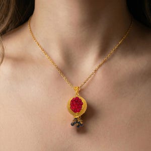 May include: A gold-colored necklace featuring a pendant shaped like a pomegranate. The pendant has a red center with a gold border and small black and blue accents at the bottom. The necklace is worn around the neck.