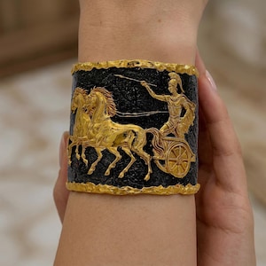 May include: Gold-toned cuff bracelet featuring a chariot scene with horses and a driver against a black background. The bracelet has a textured surface and a gold border, showcasing a classical design.