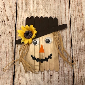 May include: A craft project of a scarecrow made from popsicle sticks. The scarecrow has a brown hat, a yellow sunflower, googly eyes, an orange triangle nose, and a black smile.