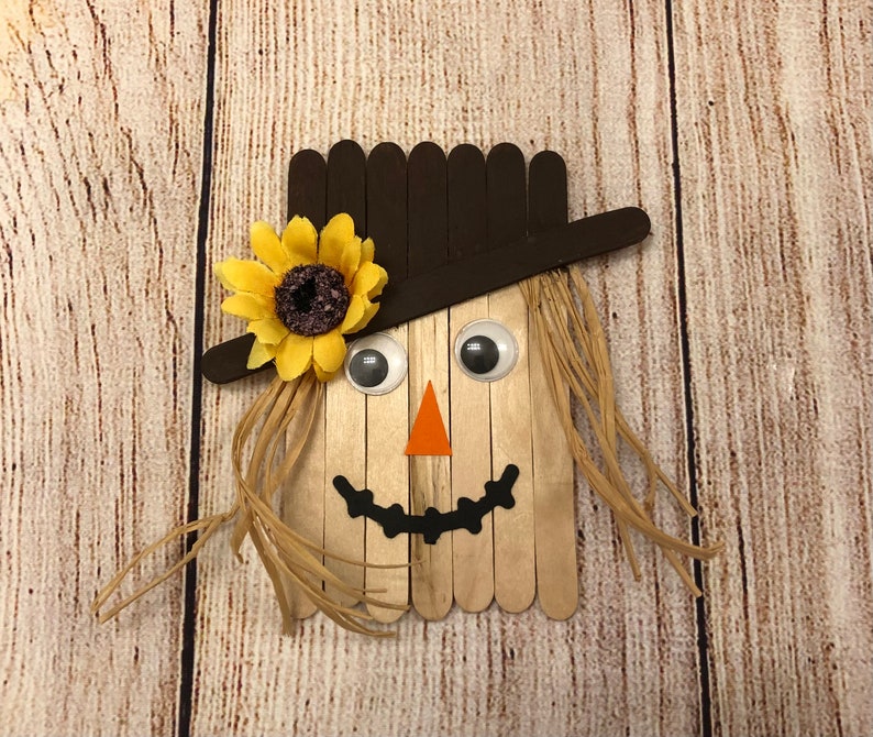 May include: A craft project of a scarecrow made from popsicle sticks. The scarecrow has a brown hat, a yellow sunflower, googly eyes, an orange triangle nose, and a black smile.