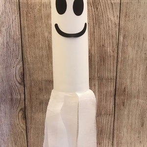 May include: A white ghost made from a cardboard tube and tissue paper. The ghost has a black smiling face with two black eyes.
