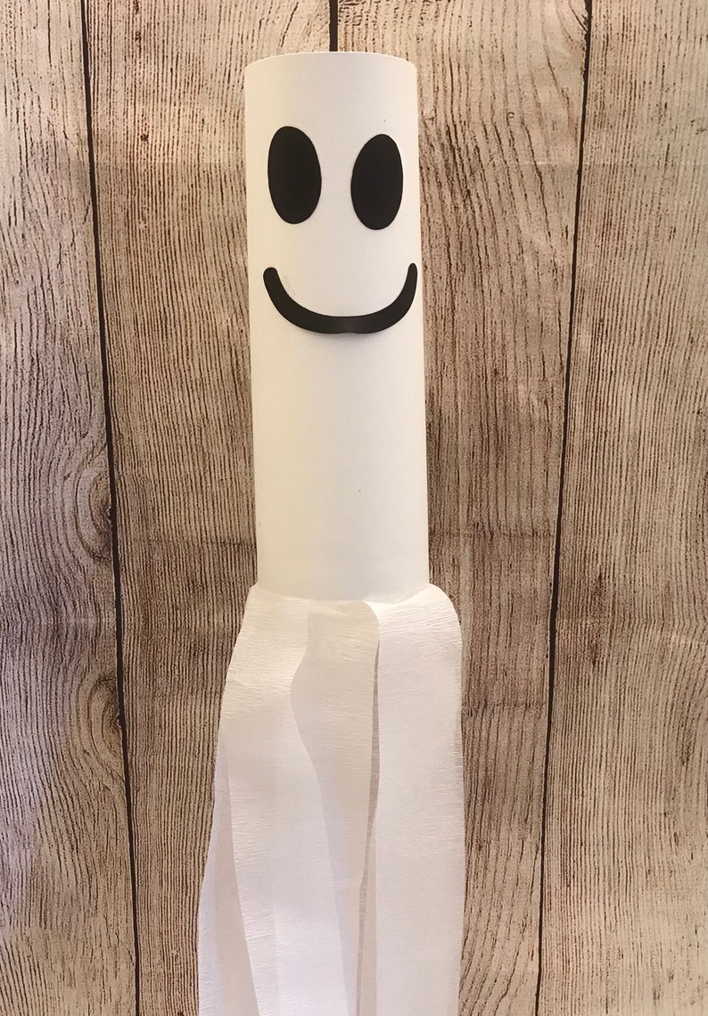May include: A white ghost made from a cardboard tube and tissue paper. The ghost has a black smiling face with two black eyes.