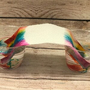 May include: A rainbow-coloured paper towel bridge connecting two clear plastic cups filled with water. The water has absorbed the colours from the paper towel, creating a colourful effect.