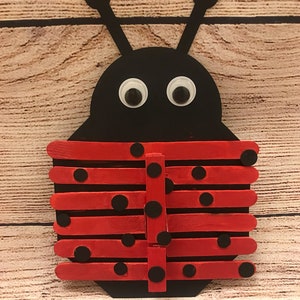 Peut inclure: Un bricolage de coccinelle rouge et noire r&eacute;alis&eacute; avec des b&acirc;tonnets de glace. La coccinelle a des points noirs sur ses ailes rouges et des yeux globuleux.