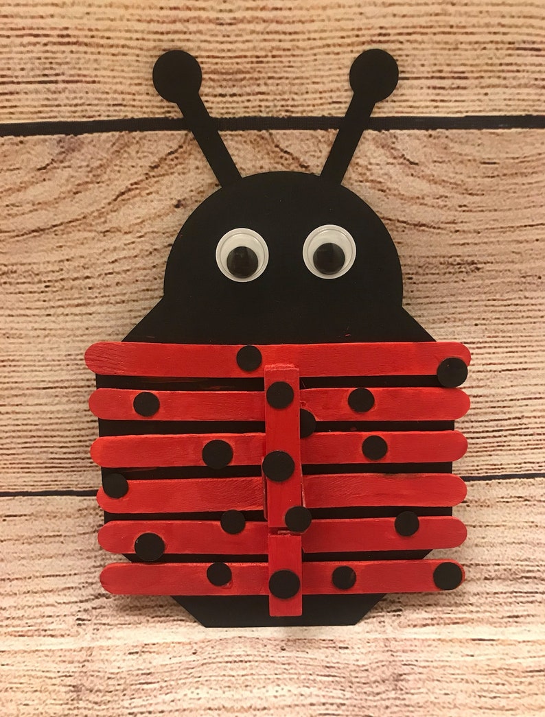 Peut inclure: Un bricolage de coccinelle rouge et noire r&eacute;alis&eacute; avec des b&acirc;tonnets de glace. La coccinelle a des points noirs sur ses ailes rouges et des yeux globuleux.