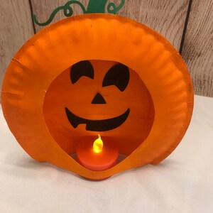 May include: A paper plate pumpkin with a carved jack-o'-lantern face and a small battery-operated tea light inside. The pumpkin is orange with a green stem and vine.