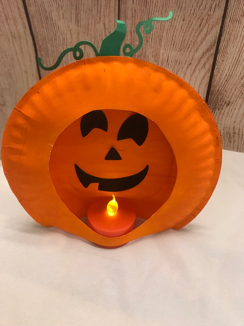 May include: A paper plate pumpkin with a carved jack-o'-lantern face and a small battery-operated tea light inside. The pumpkin is orange with a green stem and vine.