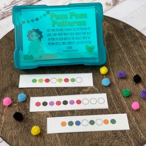May include: A children's educational game with the text "Pom Pom Patterns" on a teal plastic container. The game includes pattern cards and colorful pom-poms. The cards have colored circles to match with the pom-poms. The game is on a wooden surface.