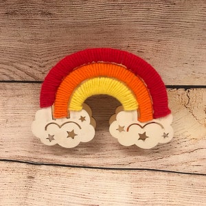 May include: A wooden rainbow with red, orange, and yellow yarn wrapped around it. The rainbow is attached to two white wooden clouds with cut-out stars.