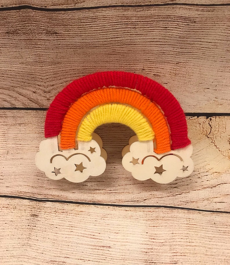 May include: A wooden rainbow with red, orange, and yellow yarn wrapped around it. The rainbow is attached to two white wooden clouds with cut-out stars.