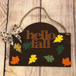 May include: A brown wooden sign with the words "hello fall" in brown letters. The sign is decorated with fall-colored leaves in shades of orange, yellow, green, and gold.