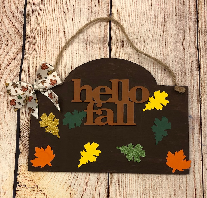 May include: A brown wooden sign with the words "hello fall" in brown letters. The sign is decorated with fall-colored leaves in shades of orange, yellow, green, and gold.