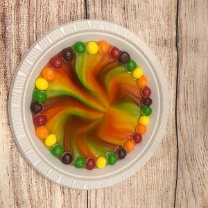 May include: A white paper plate with a swirling pattern of red, orange, yellow, and green liquid. The liquid is surrounded by a circle of colourful M&M's sweets.