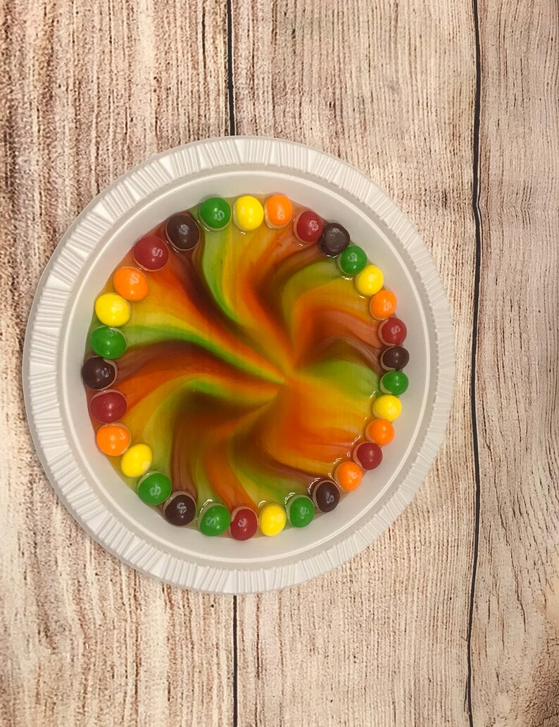 May include: A white paper plate with a swirling pattern of red, orange, yellow, and green liquid. The liquid is surrounded by a circle of colourful M&M's sweets.