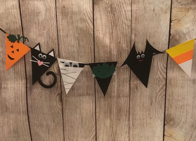 May include: A Halloween banner with six triangular flags. The flags are decorated with a pumpkin, a black cat, a mummy, a monster, a bat, and a candy corn. The flags are strung together with a white string.
