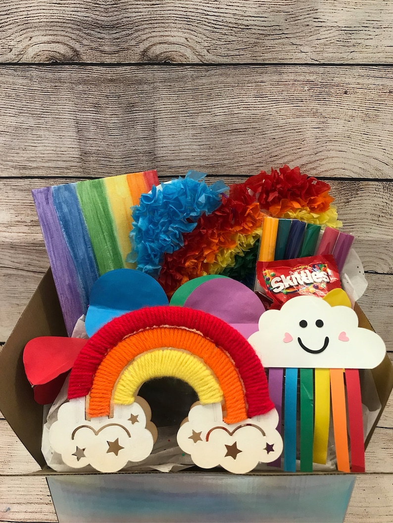 May include: A rainbow-themed craft kit in a brown box. The kit includes a rainbow yarn wrapped around a wooden rainbow, a cloud with a smiley face and rainbow streamers, tissue paper, and a bag of Skittles sweets.