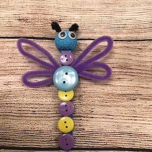Peut inclure: Une libellule artisanale de couleur violette et bleue r&eacute;alis&eacute;e avec des cure-pipes, des boutons et une boule de mousse bleue. La libellule a des yeux globuleux et des antennes en cure-pipe noires.