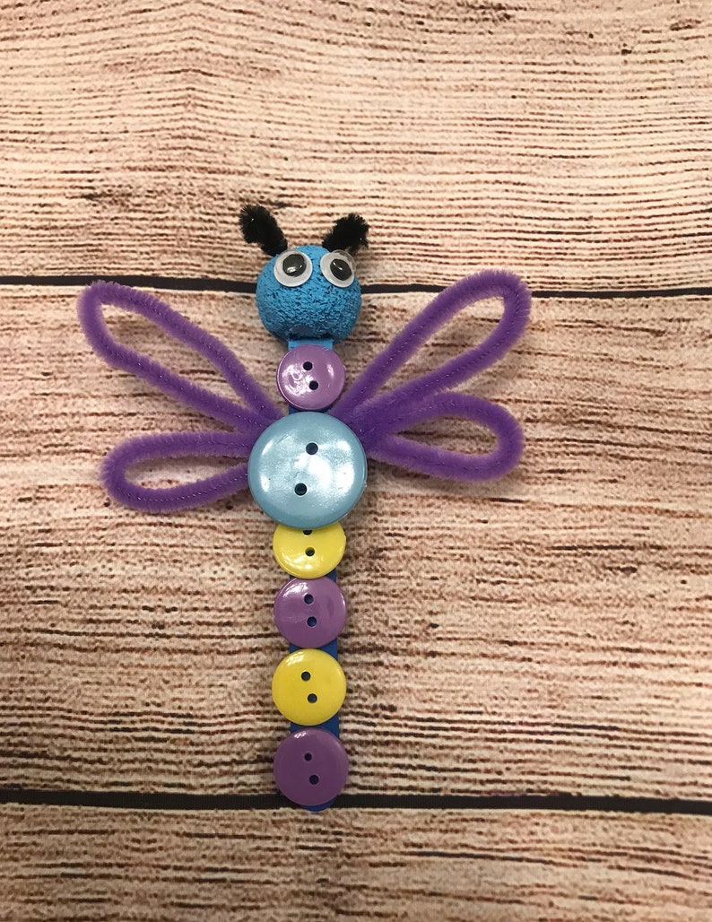 Peut inclure: Une libellule artisanale de couleur violette et bleue r&eacute;alis&eacute;e avec des cure-pipes, des boutons et une boule de mousse bleue. La libellule a des yeux globuleux et des antennes en cure-pipe noires.