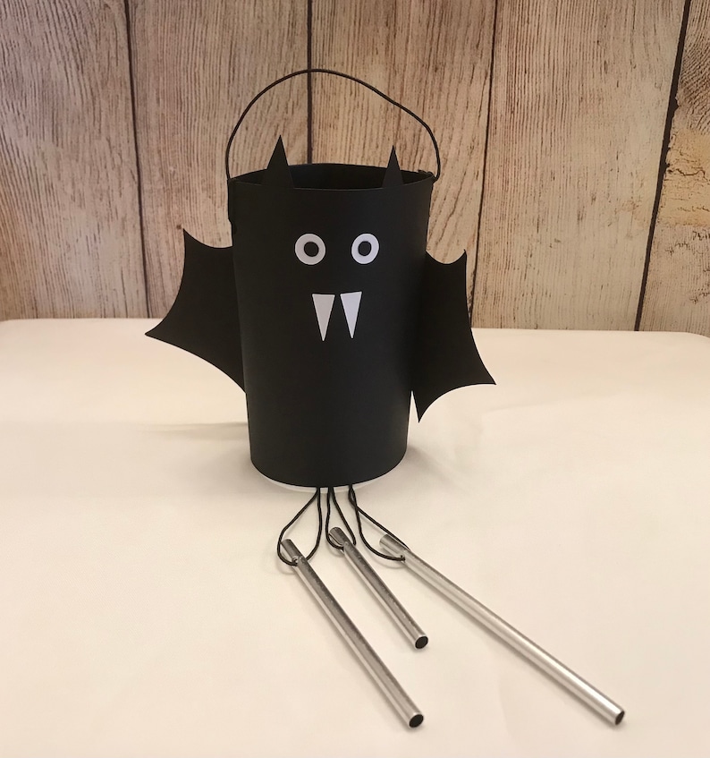 May include: Black bat-shaped wind chime with a white face and fangs. The chime has a black cylinder with a handle and three silver metal chimes hanging from it.