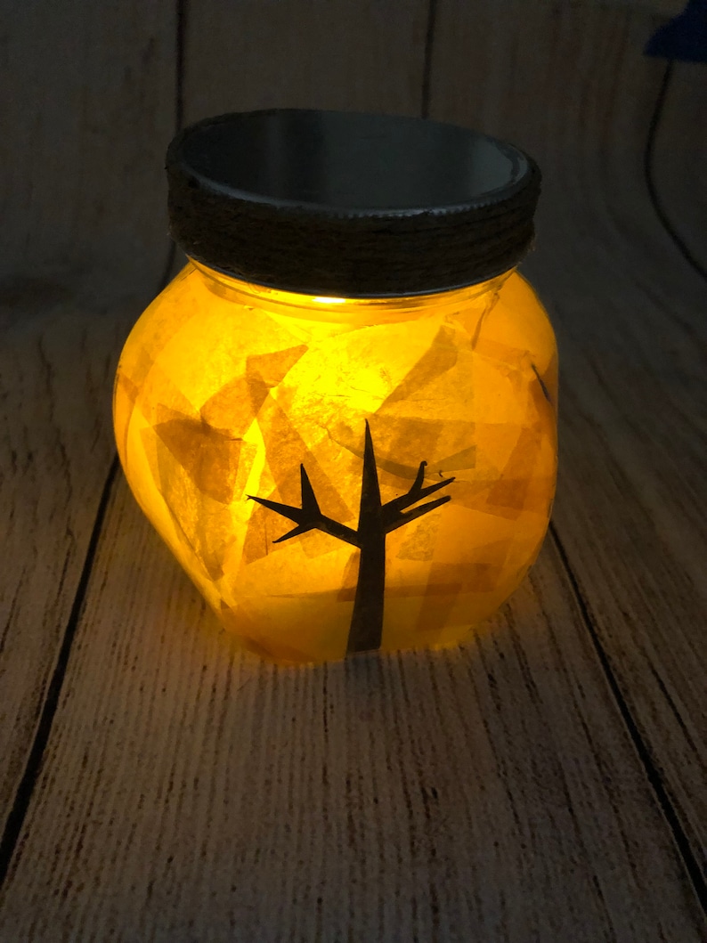 May include: A glass jar with a black silhouette of a tree inside. The jar is filled with yellow tissue paper and lit from within, creating a warm glow.