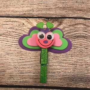 Peut inclure: Une pince &agrave; linge verte avec un motif de papillon rose et vert. Le papillon a des yeux globuleux et une bouche souriante.